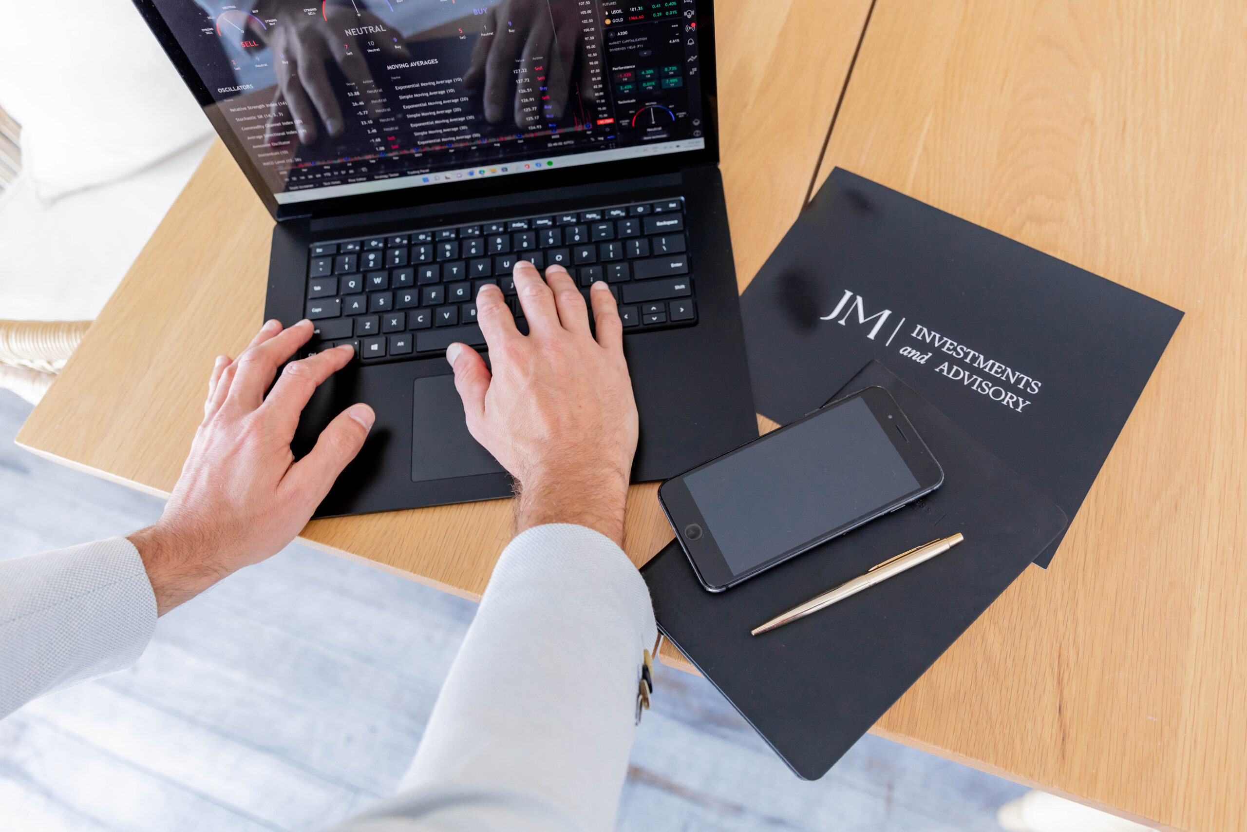 Jon Moses hands on his laptop with phone and pen on the side, JM Investments logo on the desk