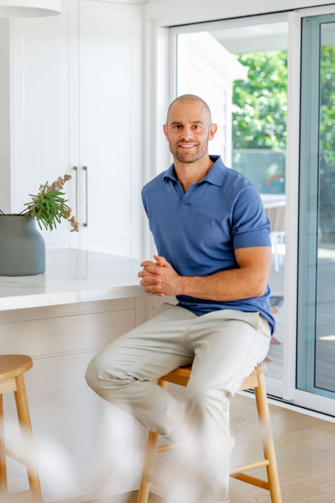 Jon Moses, Managing Director of JM Investments, sitting on a barstool in a kitchen, wearing a blue polo shirt and cream pants.